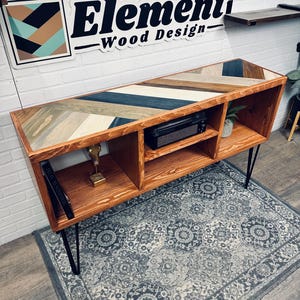 May include: A wooden console table with a chevron pattern top in shades of brown, blue, and white. The table has four open shelves and black hairpin legs. The table is sitting on a patterned rug.
