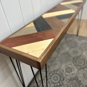 May include: A long, narrow console table featuring a rectangular top with a multi-colored herringbone pattern made of various wood tones including light, dark, and reddish-brown planks. The table is supported by four thin, black metal hairpin legs.