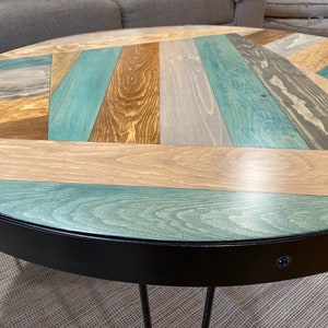 May include: Round wooden coffee table with a black metal base and a chevron pattern top made of various shades of wood, including brown, beige, and blue.