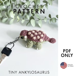 Peut inclure: Un modèle de crochet PDF pour un minuscule porte-clés Ankylosaure. Le dinosaure amigurumi présente un dos mauve foncé avec des taches rose clair, un corps vert sauge et des yeux en perles noires. Un anneau métallique avec une clé est attaché.
