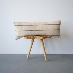 Peut inclure: Un long coussin rectangulaire de couleur crème avec des rayures brunes repose sur un tabouret en bois. Le coussin est fait d'un tissu tissé et a une texture subtile.