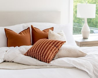 May include: A white bed with a white duvet cover and a white headboard. There are four pillows on the bed, two are brown, one is white with a pattern, and one is a beige throw pillow. The bed is in a bedroom with a white wall and a window with a view of green trees. There is a white lamp on a wooden nightstand next to the bed.