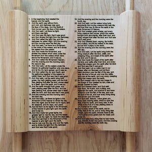 May include: A wooden scroll with text from Genesis, featuring a light-colored wood frame and wooden dowels. The text is printed in black ink, and the scroll is displayed on a neutral surface.
