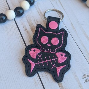 May include: Black and pink embroidered cat skull keychain with a pink fish bone design. The keychain is attached to a silver key ring.