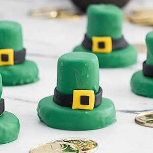Peut inclure: Plusieurs friandises vertes en forme de chapeau, avec des bandes noires et des boucles jaunes, ressemblant à des chapeaux de leprechaun. Des décorations de pièces d'or sont également présentes, évoquant la Saint-Patrick.