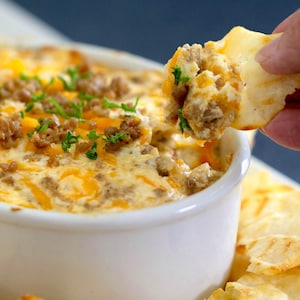 May include: A close-up shot of a white bowl filled with a creamy, cheesy dip. The dip is topped with browned sausage crumbles and shredded cheddar cheese. A piece of flatbread is dipped into the dip, showing the texture and ingredients. Fresh parsley is sprinkled on top.
