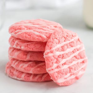 Op de afbeelding: Een stapel van vijf ronde, felroze koekjes, elk met een getextureerd oppervlak en een dunne, diagonale streep wit glazuur over de bovenkant. De koekjes zijn uniform van grootte en vorm, met één koekje leunend tegen de stapel.