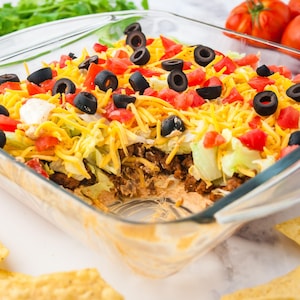 Puede incluir: Un dip en capas en un plato de vidrio transparente. El dip incluye una capa inferior de salsa cremosa, seguida de carne molida sazonada, lechuga rallada, queso cheddar rallado, tomates en cubitos y aceitunas negras en rodajas. Se ven totopos.