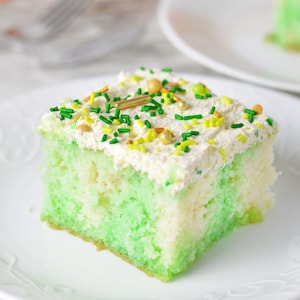 Peut inclure: Une part carrée de gâteau à couches vertes et blanches, garnie d'un glaçage blanc et de vermicelles colorés. Les vermicelles comprennent des formes vertes, jaunes et dorées. Le gâteau est posé sur une assiette blanche.