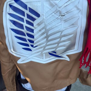 May include: Tan jacket with a large white and blue emblem on the back. The emblem is a stylized wing design with blue and white details. A red scarf is draped over the shoulder.