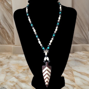 Op de afbeelding: Een lange ketting met een pijlpunthanger. De ketting heeft een kralenontwerp met witte, zwarte, zilveren en turquoise kralen. De hanger is bruin met witte chevronstrepen.
