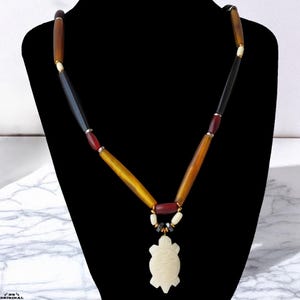 May include: A handcrafted necklace featuring a carved turtle pendant. The necklace has a mix of wooden beads in shades of brown, black, and yellow, with red and white accents. The pendant is a cream color and has carved details.