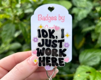 May include: White badge reel with a black and white graphic that says "IDK, I just work here". The graphic has pink hearts, yellow stars, and purple flowers. The badge reel is on a white card with the text "Badges by" on it.