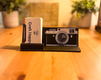 May include: A black and white illustration of a vintage camera on a wooden desk with a business card holder that says "Craft Happy" and a website address.