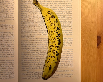 May include: A close-up of a yellow banana with brown spots, used as a bookmark in an open book. The book is about knife handling and safety. The text on the page reads "Knife-handling 401 and tricky. Why not fire-circle stone if you hold it in your hand, and the can of dog is still closed. Or that your pal's dog, will chew it up if you hold it in your hand and snap with your hip and snap"