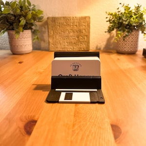 May include: A black 3D printed business card holder shaped like a floppy disk. The holder has a white business card inside with the text "3D Printing" and a logo of a 3D printer. The business card also has the text "Craft Haven" on it.