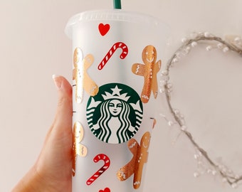 May include: A clear plastic reusable cup with a green straw and gold gingerbread men, red candy canes, and red hearts. The cup has the Starbucks logo in green and white.