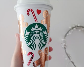 May include: White reusable coffee cup with a green Starbucks logo and gold gingerbread men, candy canes, and hearts.