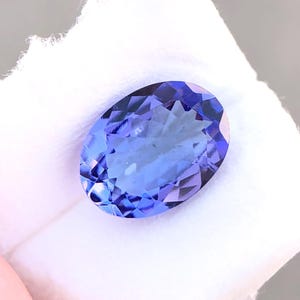 Könnte beinhalten: Ein oval geschliffener Tansanit-Edelstein mit lebendiger blau-violetter Farbe. Der Edelstein liegt auf einem weißen Hintergrund. Die Facetten erzeugen einen funkelnden Effekt, der die Klarheit und Brillanz des Steins hervorhebt. Der Edelstein ist ein Blickfang.