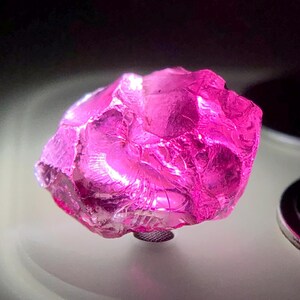Puede incluir: Una gema rosa facetada de forma irregular. La gema es de un color magenta vibrante, con una estructura cristalina. La piedra está montada sobre una pequeña base plateada. La iluminación resalta la estructura interna y el color de la gema.