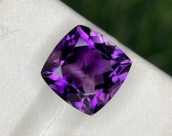 Könnte beinhalten: Ein tiefvioletter Amethyst-Edelstein mit einem Kissen-Schliff. Der Edelstein ist facettiert und hat eine hohe Klarheit.