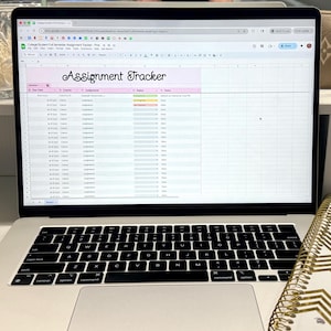 May include: A laptop displaying a spreadsheet titled "Assignment Tracker" with a pink header. A spiral notebook with gold geometric designs and a keyboard are in the foreground. Other items are on a white shelf.