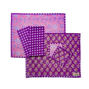 Puede incluir: Un conjunto de mantelería que incluye un mantel individual rectangular con estampado floral morado y blanco, un posavasos cuadrado a juego, dos servilletas de cuadros morados y blancos y un tapete rectangular con estampado de conchas.