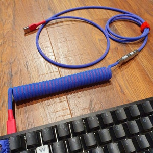 Könnte beinhalten: Ein gewickeltes blaues und pinkfarbenes USB-Kabel mit einem roten Stecker und einem silbernen Stecker. Das Kabel ist gewickelt und liegt auf einer schwarzen Tastatur mit weißen Tasten, die mit Zahlen und Symbolen beschriftet sind.