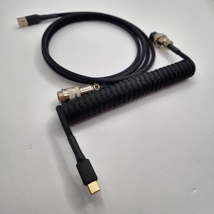 Könnte beinhalten: Ein schwarzes, gewickeltes Kabel mit einem USB-C-Stecker an einem Ende und einem vergoldeten Stecker am anderen Ende.