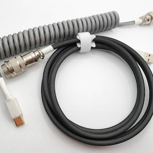 Puede incluir: Un cable gris y blanco en espiral con un conector plateado y un cable negro con un conector USB. El cable está diseñado para teclados mecánicos y otros dispositivos electrónicos. El cable está sobre un fondo blanco.