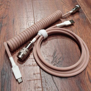 May include: A coiled cable with a white and beige braided outer covering. The cable has two metal connectors, one on each end. The cable is coiled and lying on a wooden surface.