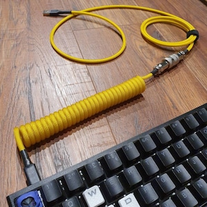 Op de afbeelding: Een gele gekrulde kabel met een USB-connector aan het ene uiteinde en een 3,5 mm-aansluiting aan het andere uiteinde. De kabel is gekruld en ligt op een houten oppervlak. Een zwart toetsenbord is op de achtergrond.