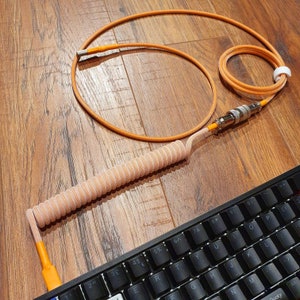 May include: A coiled orange and white cable with a black and white keyboard in the background. The cable has a coiled section and a straight section with a connector on the end.