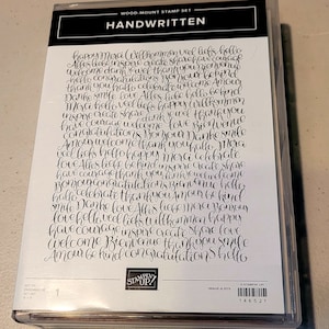 May include: A clear plastic case containing a wood-mount stamp set. The stamp set is labeled "HANDWRITTEN" and features a collection of words in a handwritten style, including "happy," "welcome," and "thank you."