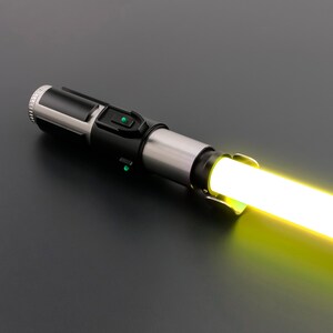 May include: A black and silver lightsaber with a bright yellow blade. The hilt features a black grip section with green activation buttons and a silver midsection. The blade emits a bright, glowing yellow light.