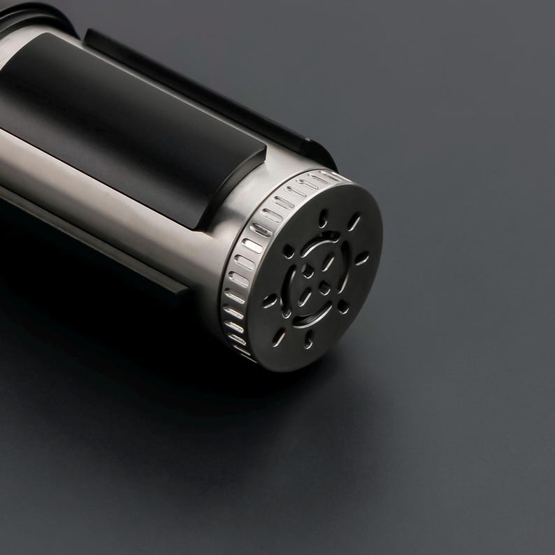 May include: A close-up of a metallic, cylindrical object with a black rectangular section. The object has a silver, circular end with multiple small holes. The background is a dark gray.
