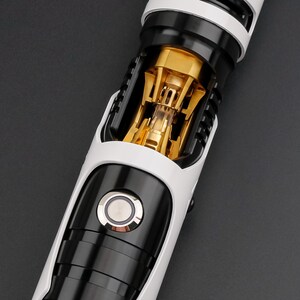 May include: A close-up of a lightsaber hilt with a white and black exterior. The hilt features a gold-colored internal mechanism visible through a clear section. A circular button is centered on the black section.