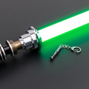 May include: A green lightsaber keychain with a silver handle and a black and silver base. The lightsaber is lit up and the keychain is attached to a silver ring.