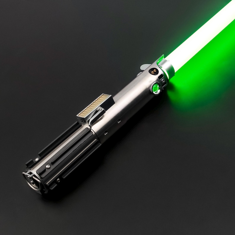 May include: A silver and black lightsaber with a bright green blade. The hilt features detailed metallic components, including buttons and a rectangular section. The glowing green blade extends from the top, creating a striking contrast against the dark background.