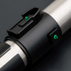 May include: Close-up of a lightsaber hilt with a black grip and a silver blade section. The hilt features a black control panel with a green light and a second green light on the side. The design is sleek and futuristic.