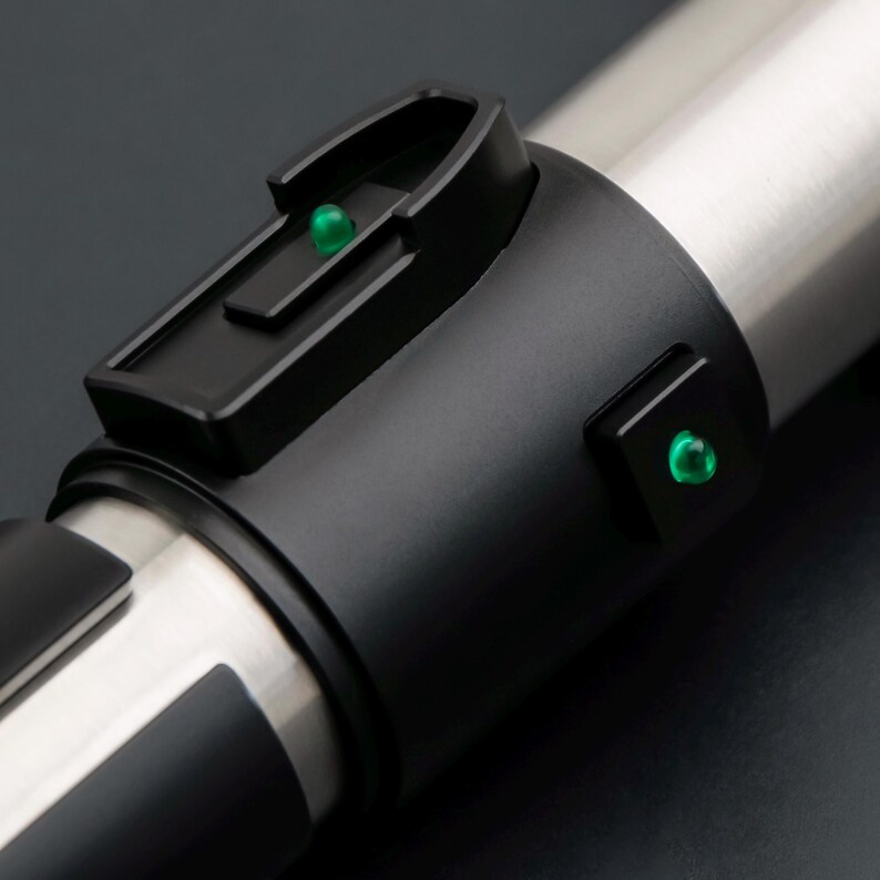 May include: Close-up of a lightsaber hilt with a black grip and a silver blade section. The hilt features a black control panel with a green light and a second green light on the side. The design is sleek and futuristic.