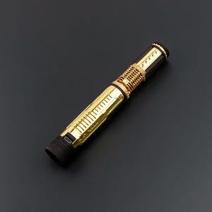 Puede incluir: Un sable de luz dorado y negro con detalles intrincados. El mango presenta un dise&ntilde;o acanalado y una serie de detalles dorados. El sable de luz tiene una est&eacute;tica elegante y futurista, con un puerto de carga visible cerca de la base. Mide unos 25 cm.