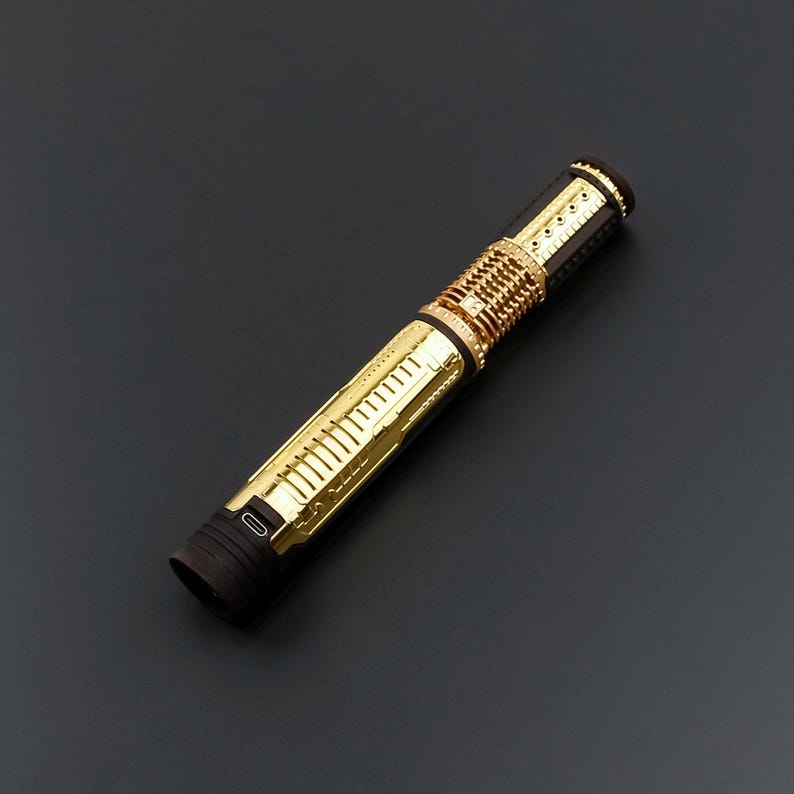 Puede incluir: Un sable de luz dorado y negro con detalles intrincados. El mango presenta un dise&ntilde;o acanalado y una serie de detalles dorados. El sable de luz tiene una est&eacute;tica elegante y futurista, con un puerto de carga visible cerca de la base. Mide unos 25 cm.