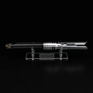 K&ouml;nnte beinhalten: Ein silberner und schwarzer Lichtschwert-Requisit auf einem klaren Acryl-St&auml;nder. Das Lichtschwert hat einen strukturierten schwarzen Griff und eine silberne Klinge.
