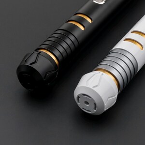 May include: Two futuristic light sabers. One is black with gold accents, the other is white with silver and gold accents. Both have ribbed handles and detailed end caps. The background is a dark gray.
