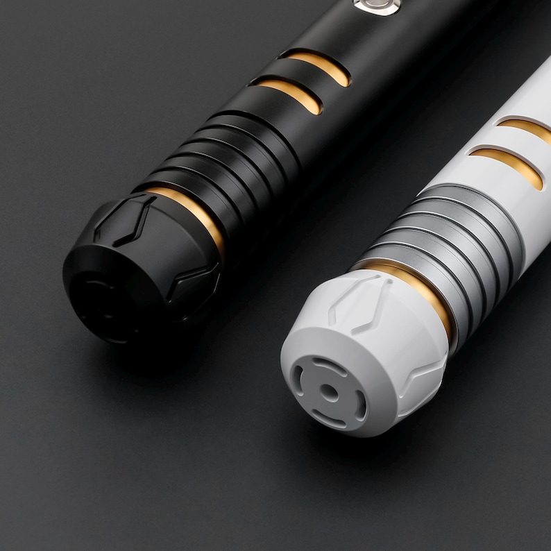 May include: Two futuristic light sabers. One is black with gold accents, the other is white with silver and gold accents. Both have ribbed handles and detailed end caps. The background is a dark gray.