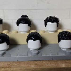 May include: A collection of Lego minifigure heads with various black hairstyles. The heads are white with different black hair styles, including a bun, short cuts, and curly styles. They are arranged on a light tan and gray Lego base.