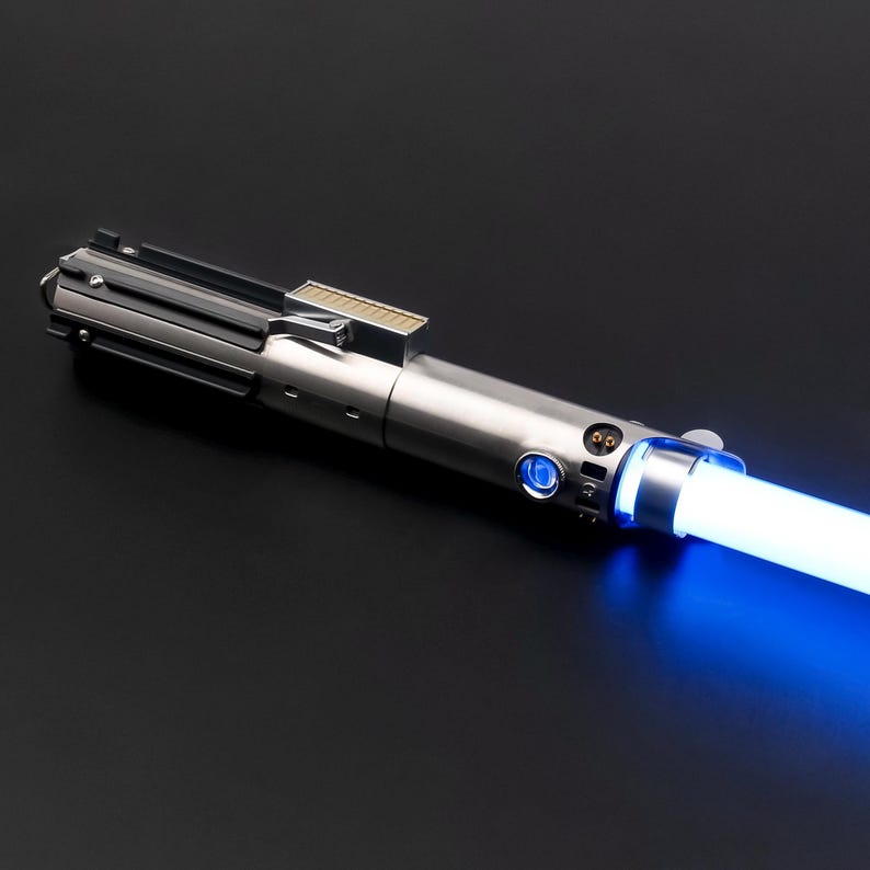 May include: A silver lightsaber with a blue glowing blade. The hilt has a metallic finish with detailed mechanical elements. The blade emits a bright, consistent blue light, suggesting it is powered on. The lightsaber is set against a dark background.