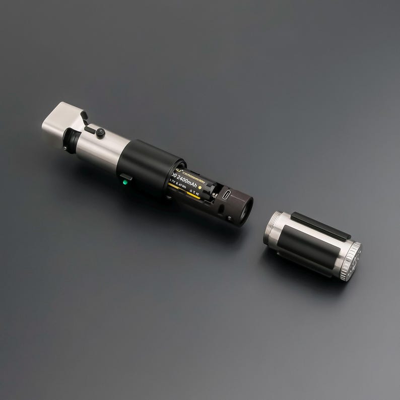 May include: A disassembled lightsaber prop with a black and silver handle. The handle is partially open, revealing the battery compartment with a 2400mAh battery. The detached end cap is also silver and black.
