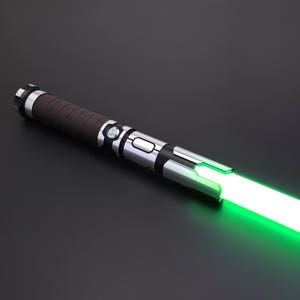 May include: A silver and black lightsaber with a brown handle and a bright green blade. The lightsaber is angled diagonally across a dark surface, with the green blade illuminated.